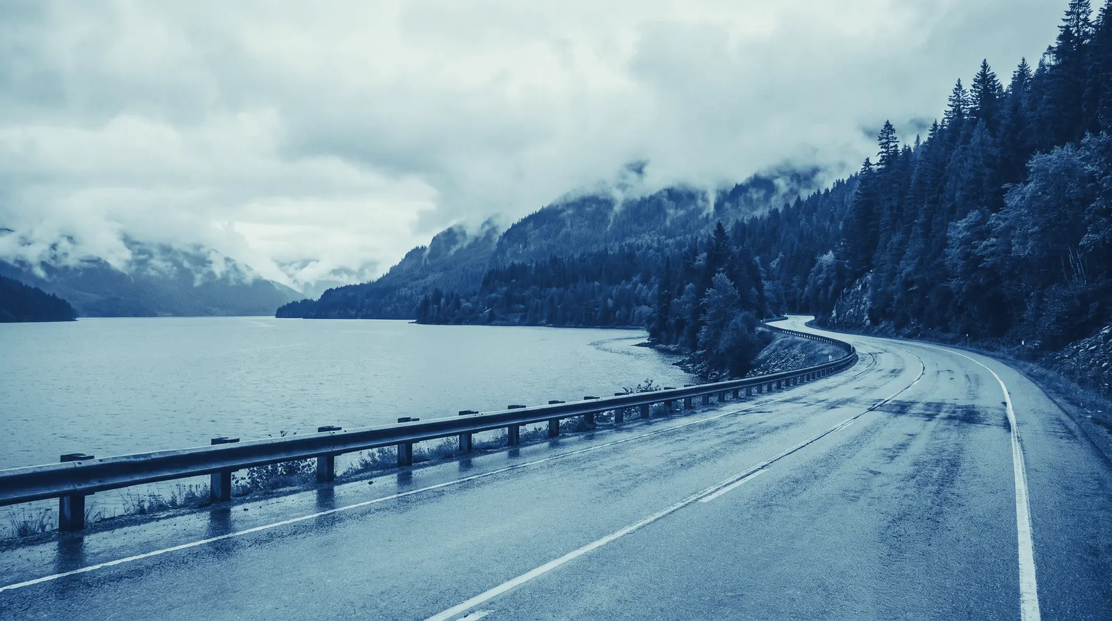 Rendering of a two-lane highway curving along a grey mountain lake under low cloud, forest on both shores