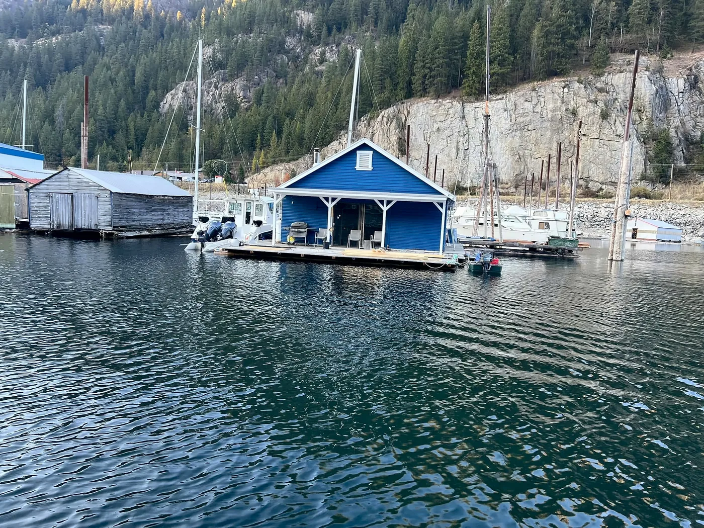 The blue floating cabin seen from the water, tied at the end of its own dock with the rock bluff behind it